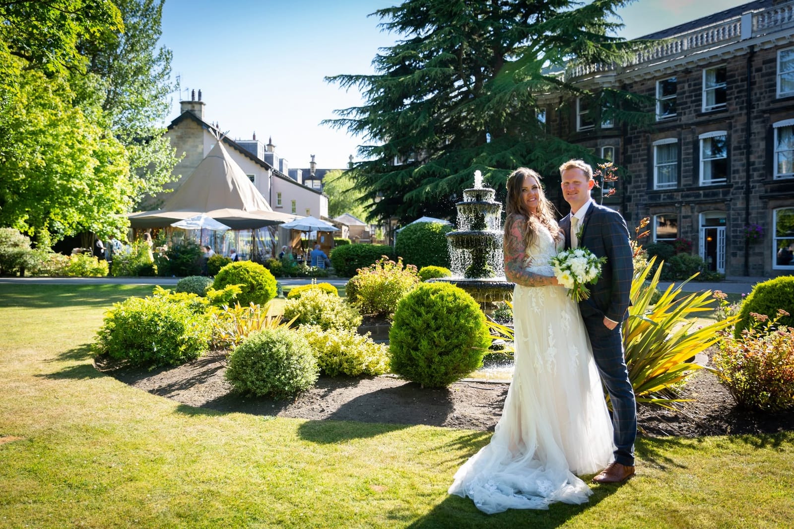 A bride and groom beside a fountain in a sunlit garden