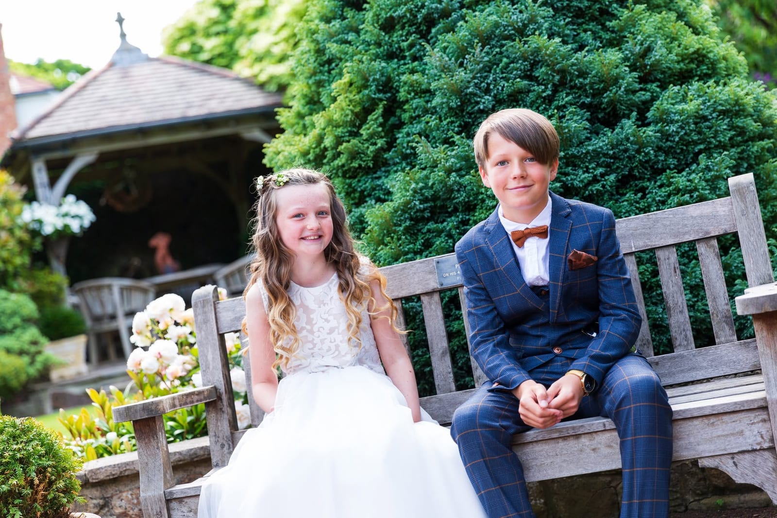 Two children dressed for a wedding, sitting together on a garden bench