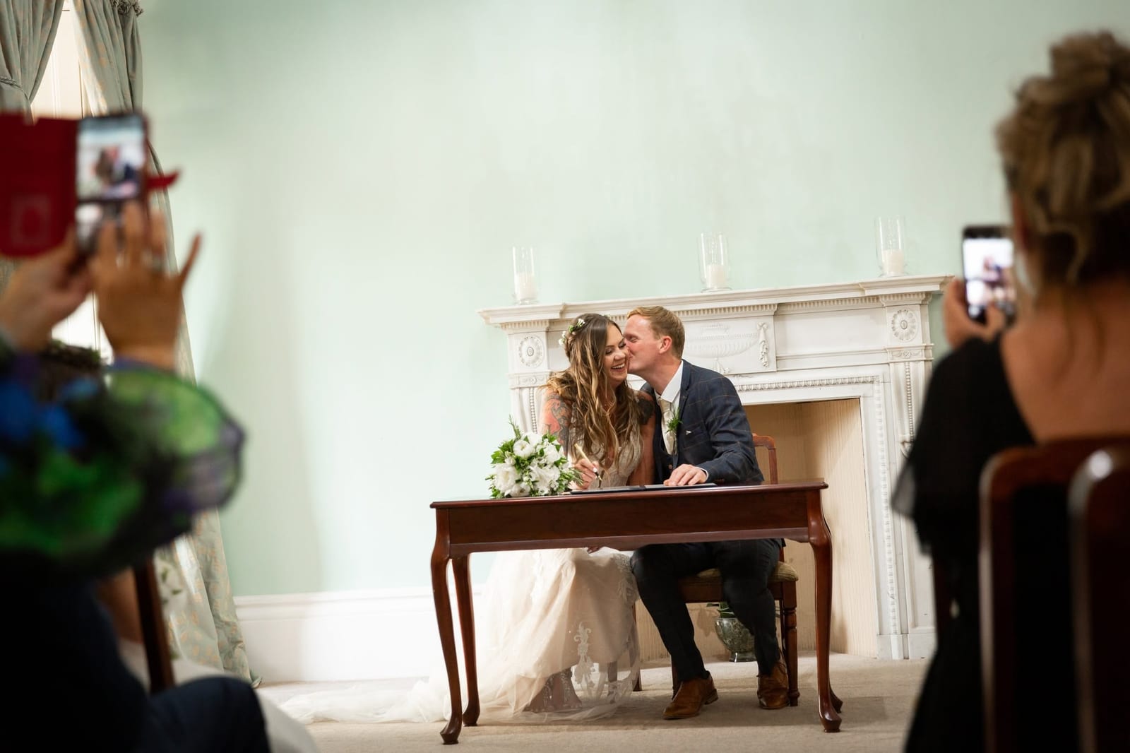 A groom kisses the bride at the signing table, framed by their guests